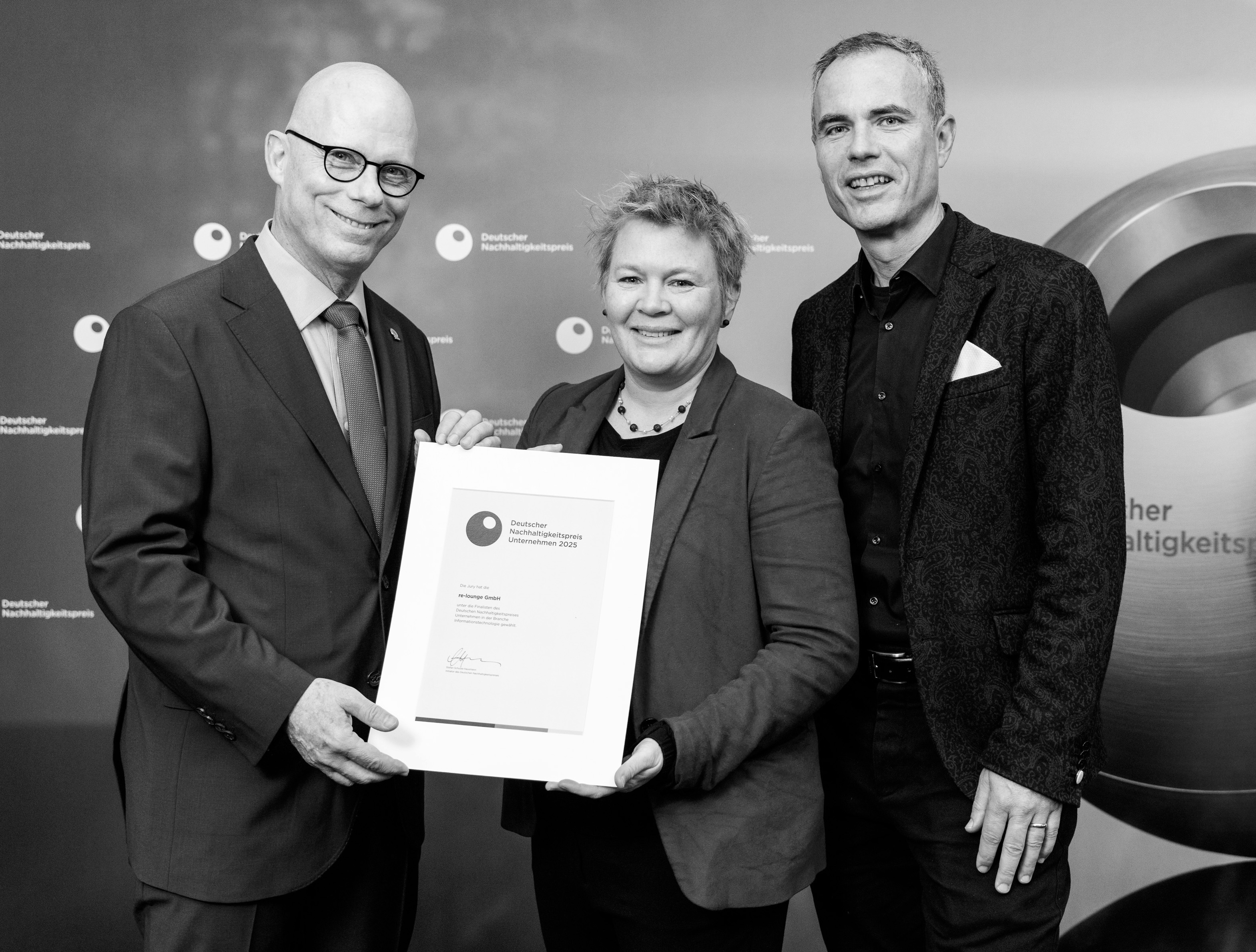 Katrin in der Mitte hält eine Urkunde, die sie gemeinsam mit zwei Männern präsentiert.