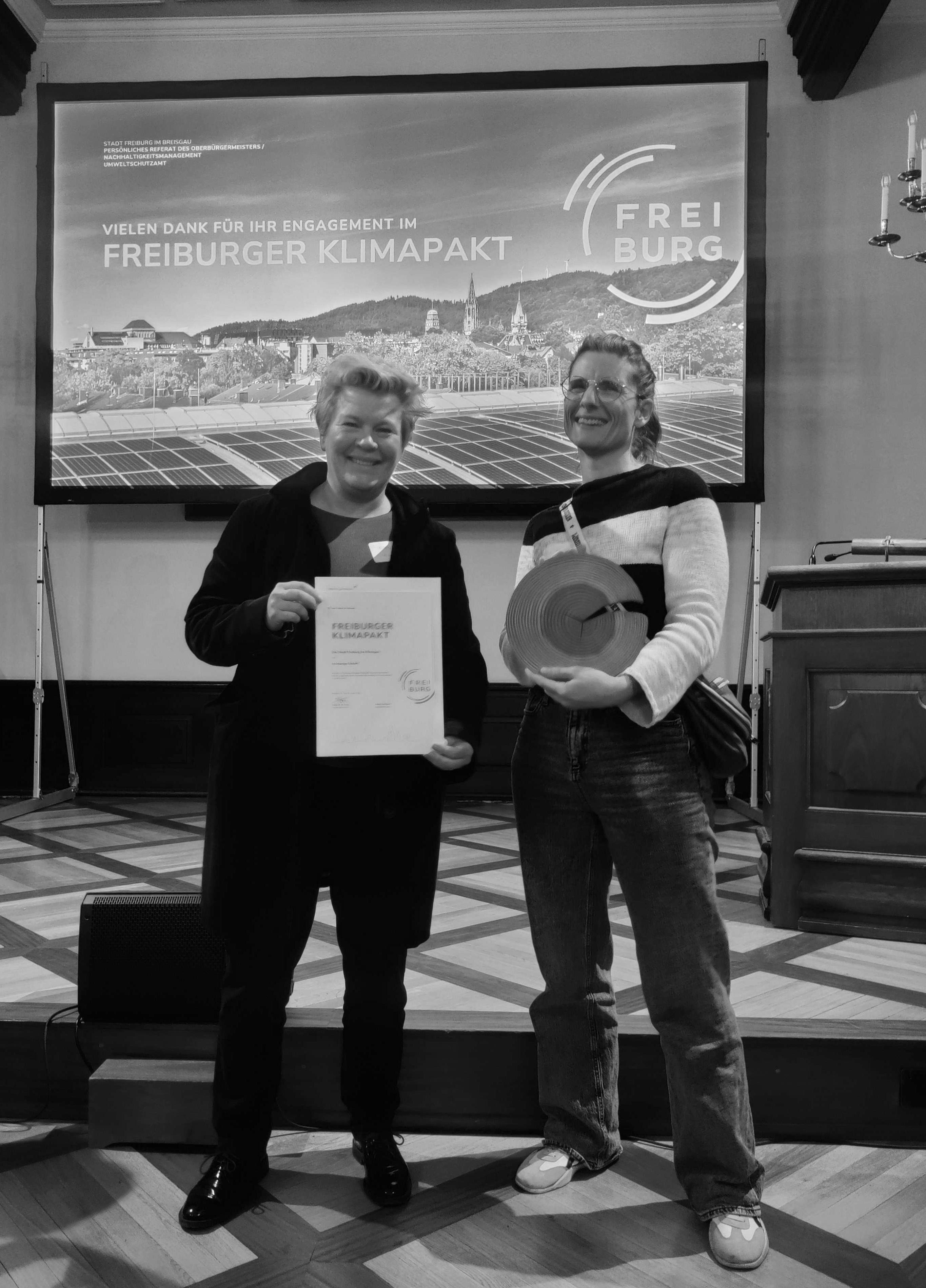 Katrin und Elli bei der Auszeichnung für unsere Teilnahme am Freiburger Klimapakt: mit Urkunde und Holztrophäe in den Händen, im Hintergrund ein Bildschirm mit Dankesbotschaft.